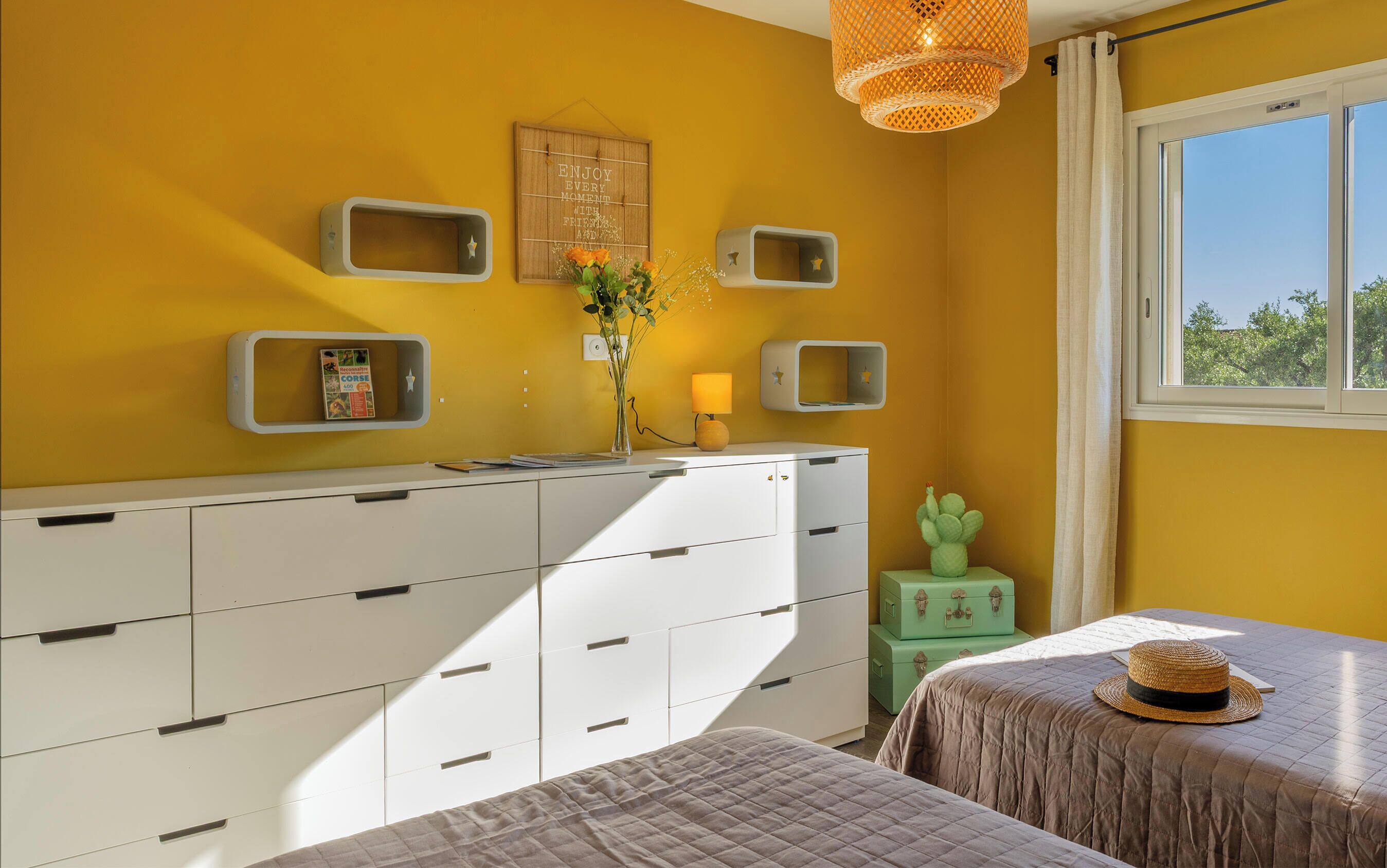 A room with a yellow wall, a white dresser with multiple drawers, a small green stool, a bed with a brown quilt, and a window with a view of trees.