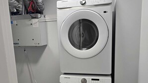 Ett tvättrum med två staplade tvättmaskiner från Samsung, en ventilationsöppning i taket och en hylla med rengöringsmedel.