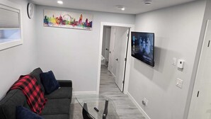 Ett modernt vardagsrum med en soffa, ett soffbord i glas, en tavla som föreställer en stadsbild, en tv och en hall som leder till ett annat rum.