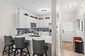 A modern kitchen with white cabinets, a central island, bar stools, and a microwave. - Heron's Watch Way 3304 (Santa Rosa Beach)