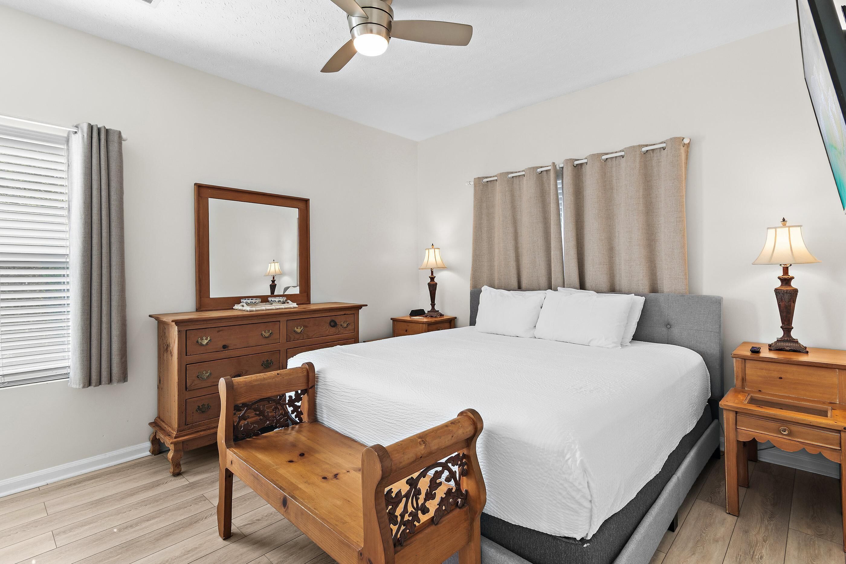 A bedroom with a bed, wooden nightstands, a dresser, a chair, a ceiling fan, and a window with curtains.