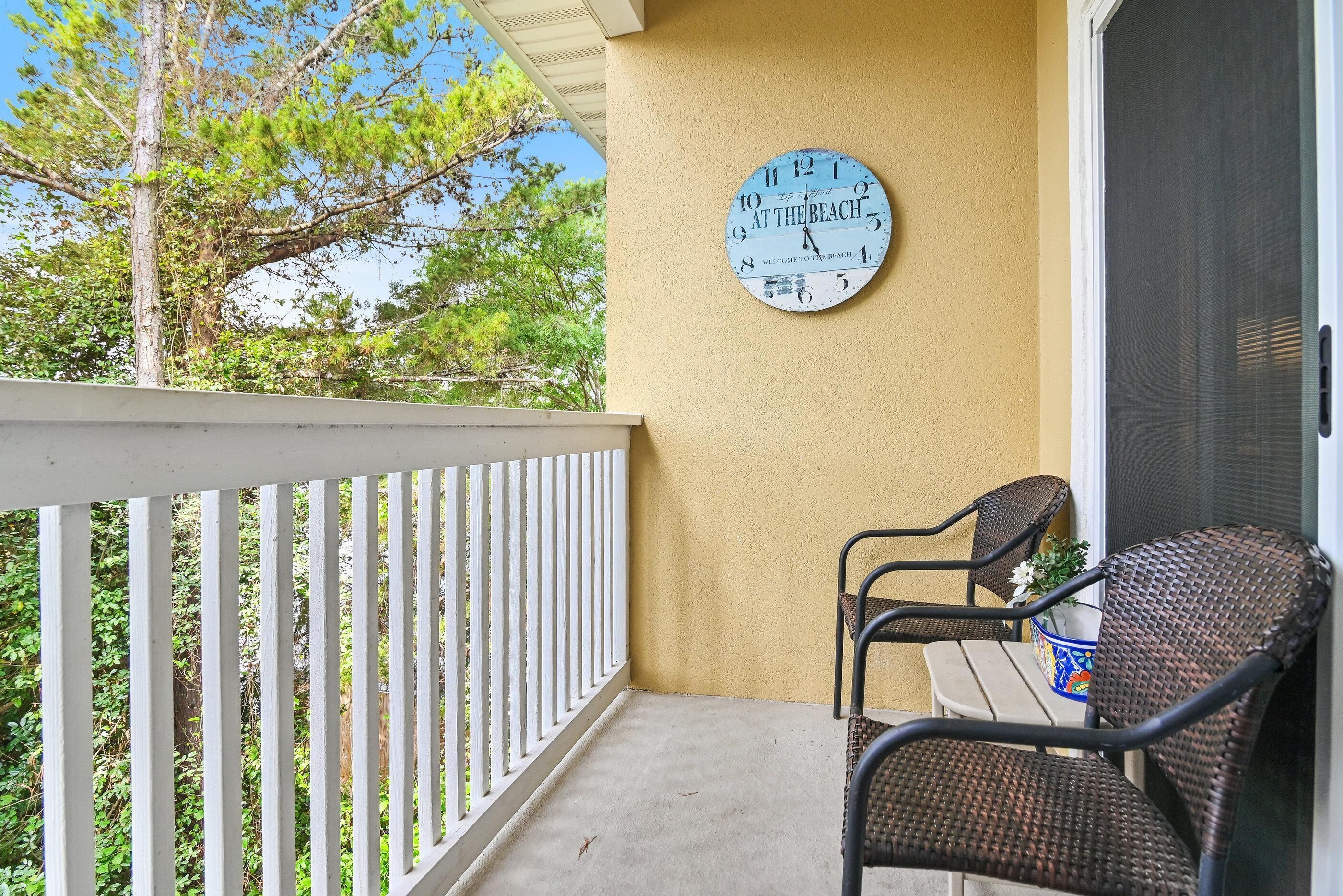 A balcony with a white railing, two wicker chairs, a table with a potted plant, and a clock on the wall.