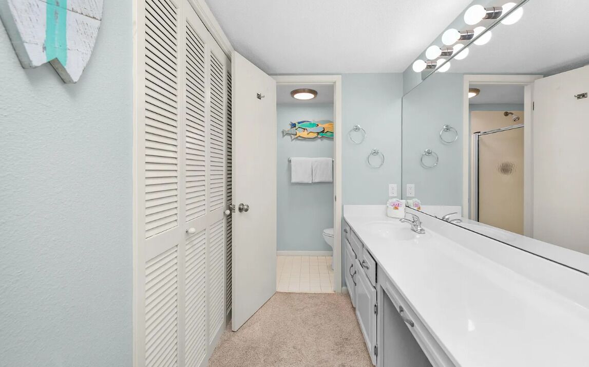 A bathroom with a white vanity, a large mirror with lights, a towel rack, and a shower area.
