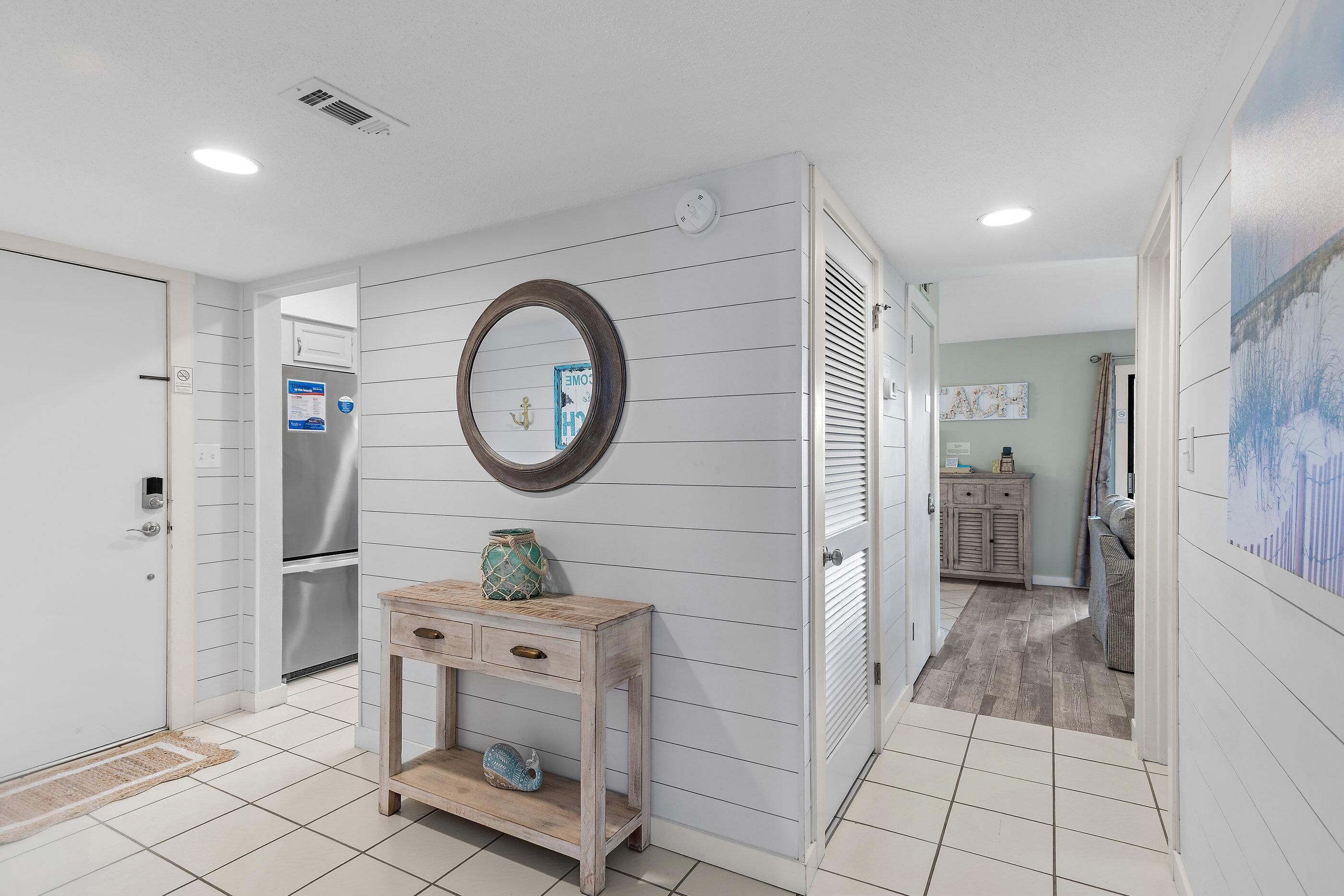 A hallway with a wooden console table, a mirror, a refrigerator, and a door with a doormat.