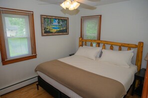 A bedroom with a bed, two windows with blinds, a ceiling fan, and a painting on the wall.