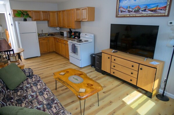 A compact kitchen with wooden cabinets, a white refrigerator, a microwave, a stove, and a large flat-screen TV.
