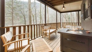 A covered outdoor deck with wooden furniture, a fire feature, and a view of trees. - Updated Chimney Rock cabin - Views & River Sounds (Chimney Rock)
