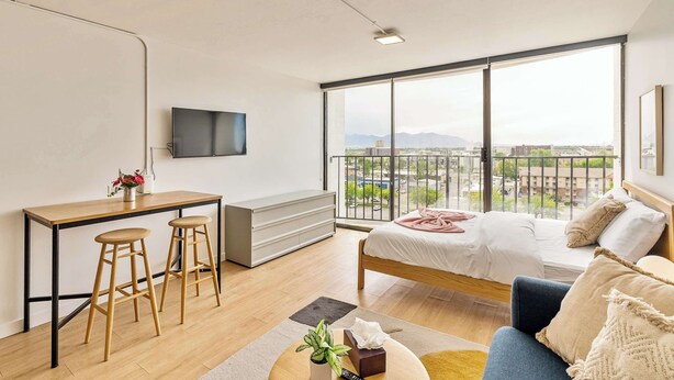 Ein modernes Schlafzimmer mit großem Fenster, einem Bett, einer Couch, einem Schreibtisch mit Hocker und einem Fernseher.