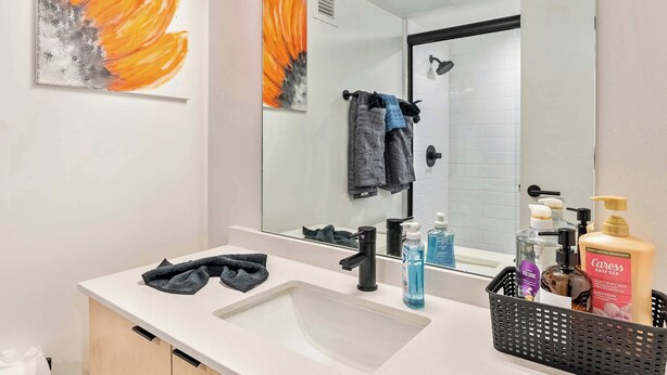 A modern bathroom with a large mirror, a white sink, and a black faucet. There are towels hanging on the wall and a basket with toiletries. - Mountain View 9th Floor City Views Free Pkg, Gym (Salt Lake City)