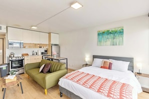 Una habitación de hotel moderna con kitchenette, un sofá verde, una cama grande con manta estampada y un cuadro en la pared.