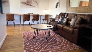 Una sala moderna con un sofá de cuero marrón, una mesa de centro redonda, una alfombra estampada y un comedor con una mesa y sillas de madera.