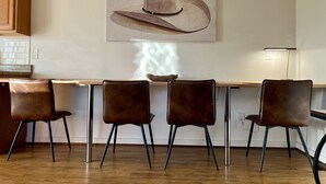Un comedor con una mesa de madera, tres sillas marrones, un cuadro colgado en la pared de un sombrero de vaquero y una lámpara.
