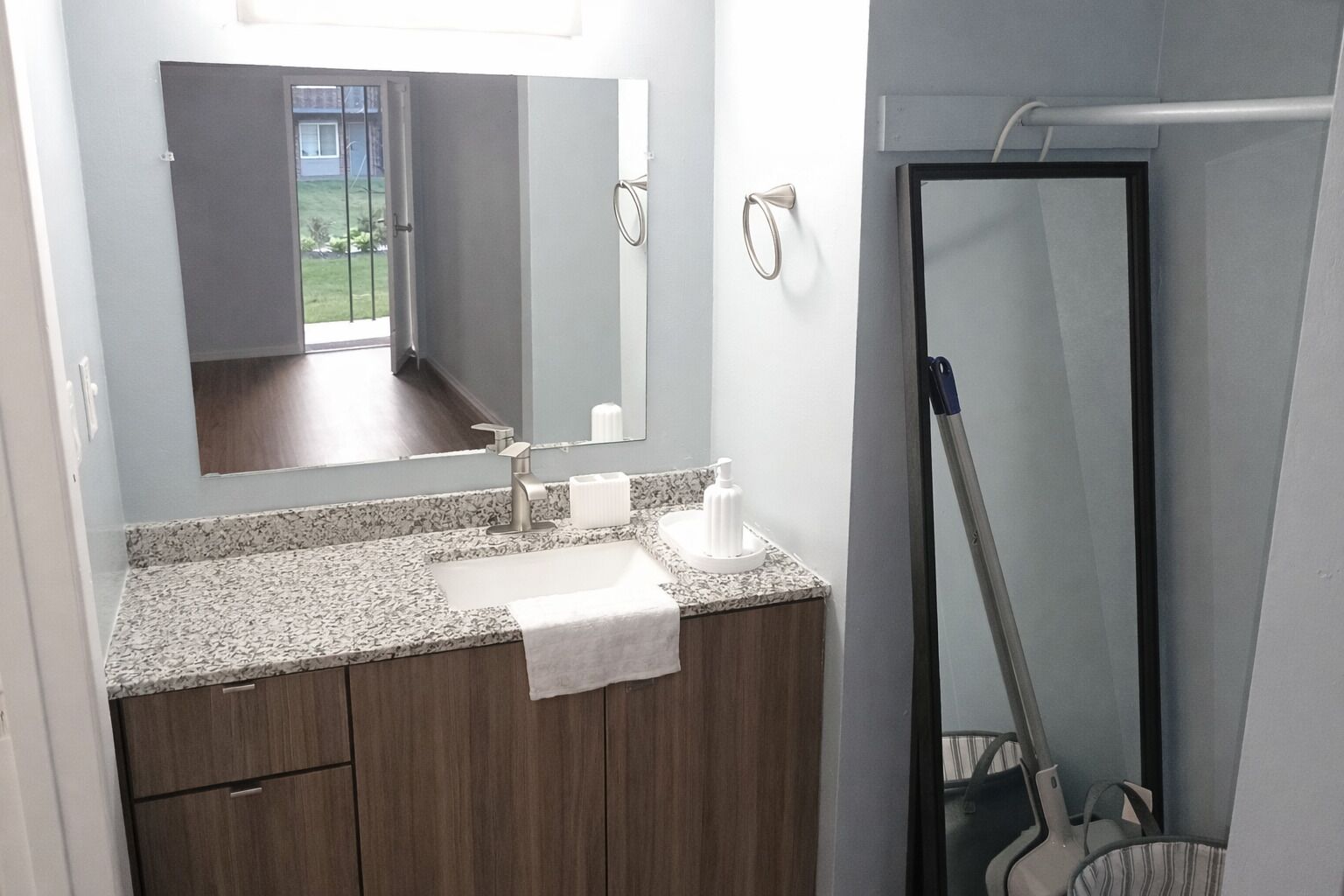 Un baño con encimera de granito, un lavabo, un espejo, una toalla y un trapeador apoyado en la pared.