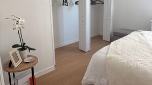 Een slaapkamer met een bed, een kast met kleding, een kleine tafel met een plant en een fotolijst.