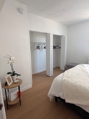 Ein Schlafzimmer mit einem Bett, einem Kleiderschrank mit Kleidung, einem kleinen Tisch mit einer Pflanze und einem Bilderrahmen.