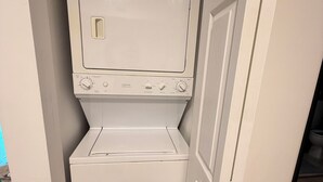 Een kleine wasruimte met een wasmachine en droger, een deur naar een andere ruimte, en een blauwe tas bovenop de droger.