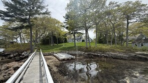 Eine Holzstele führt zu einer felsigen Uferlinie mit einem Teich, umgeben von Bäumen und Häusern im Hintergrund.