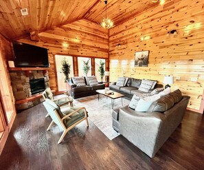 A log cabin interior with a stone fireplace, a television mounted on the wall, and a large window. - Luxury Lodge-Private Chef-Private Heated Pool-Amazing Views-Sleeps 24 (Sevierville)