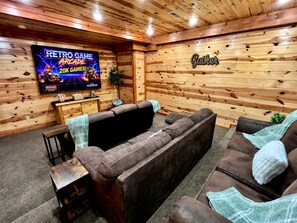 Une pièce chaleureuse avec un grand téléviseur à écran plat diffusant un jeu vidéo rétro, un mur en bois orné d’une inscription « Rassemblement », et un canapé en cuir marron.