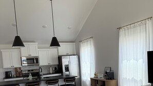 A modern kitchen with white cabinets, a microwave, and a refrigerator. There is a dining area with a bar stool and a sofa.