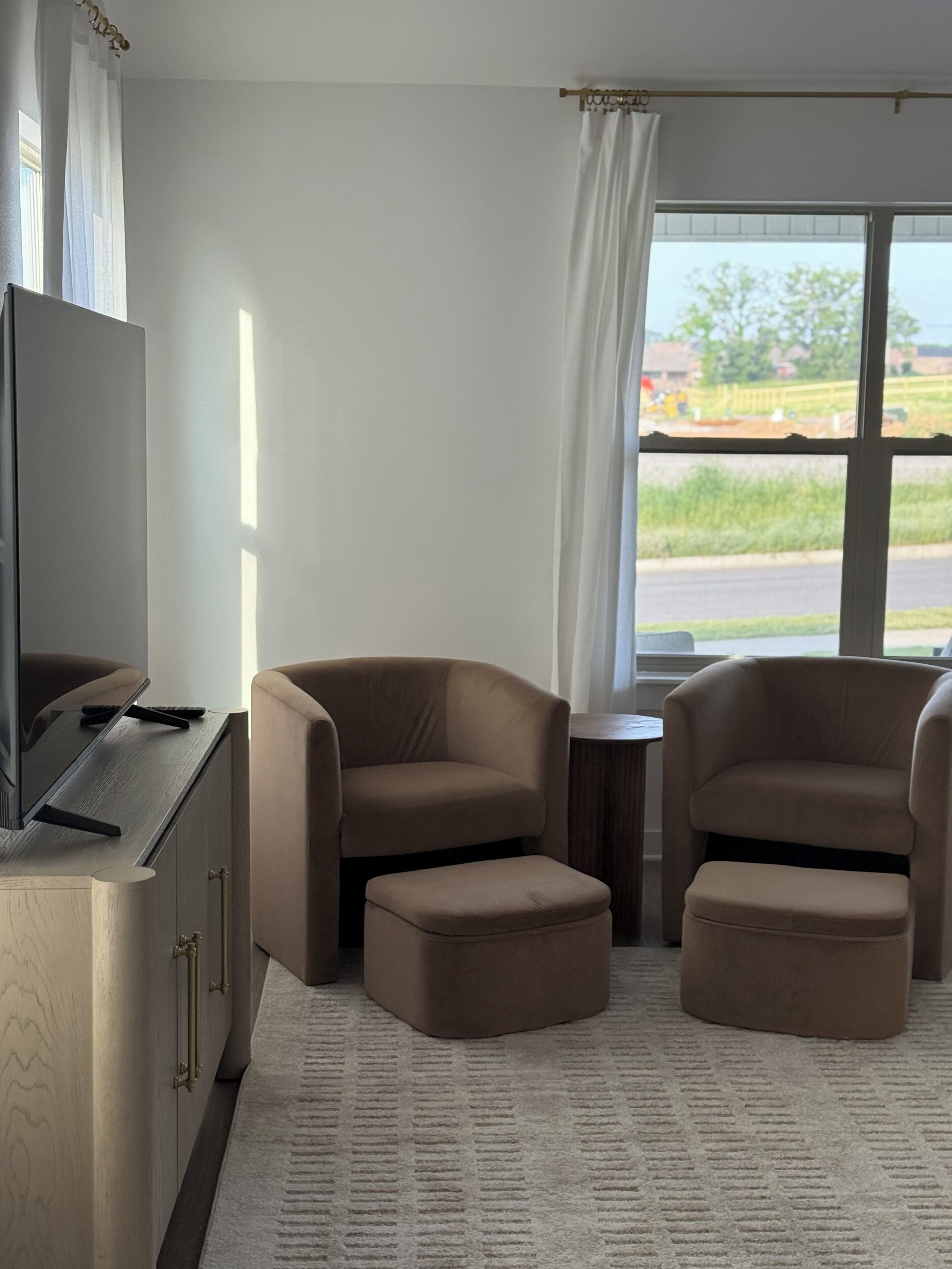 Una habitación con una televisión de pantalla plana, dos sillones marrones, un pequeño reposapiés, una mesita de madera y un ventanal con vistas a la vegetación y una carretera.