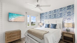 Une chambre à coucher comprenant un lit, un ventilateur de plafond, un téléviseur, une fenêtre et un mur orné d’un motif floral.