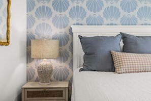Ein Schlafzimmer mit einem weißen Bett, einem grauen Kissen, einem karierten Kissen, einer Lampe und einer Wand mit Muschelmuster.