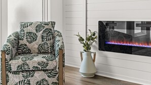 Une pièce avec un plancher en bois, une chaise ornée d’un motif de feuilles, un vase blanc contenant des plantes et une cheminée avec un feu illuminé de bleu.
