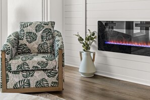 A room with a wooden floor, a chair with a leaf pattern, a white vase with plants, and a fireplace with a blue-lit fire. - Brand NEW Luxury Beachfront Paradise ~ Minutes to 30a! (Panama City Beach)