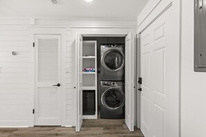 A laundry room with two stacked front-loading washers and dryers, a white door with a handle, and a shelf with laundry supplies. - Brand NEW Luxury Beachfront Paradise ~ Minutes to 30a! (Panama City Beach)