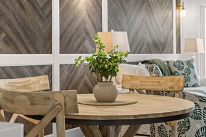 Ein runder Holztisch mit einer Vase grüner Pflanzen, umgeben von Holzstühlen, in einem Raum mit Wänden im Fischgrätmuster und einem Bett mit gemusterten Betttextilien.