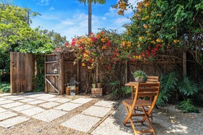 Un patio con mesa y sillas de madera, rodeado de un jardín con plantas en flor y una valla de madera.