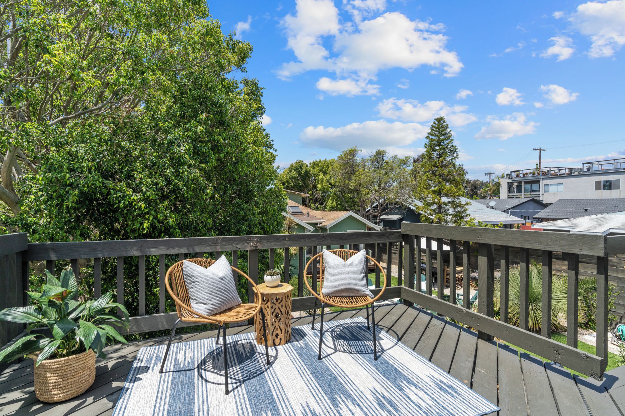A wooden deck with two wicker chairs and a small table, surrounded by lush greenery and a clear sky.