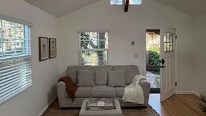 A spacious living room with a sofa, ottoman, and a large window with blinds. - Newly renovated, Stylish Studio Beach Bungalow a couple of blocks from the beach (Los Angeles)