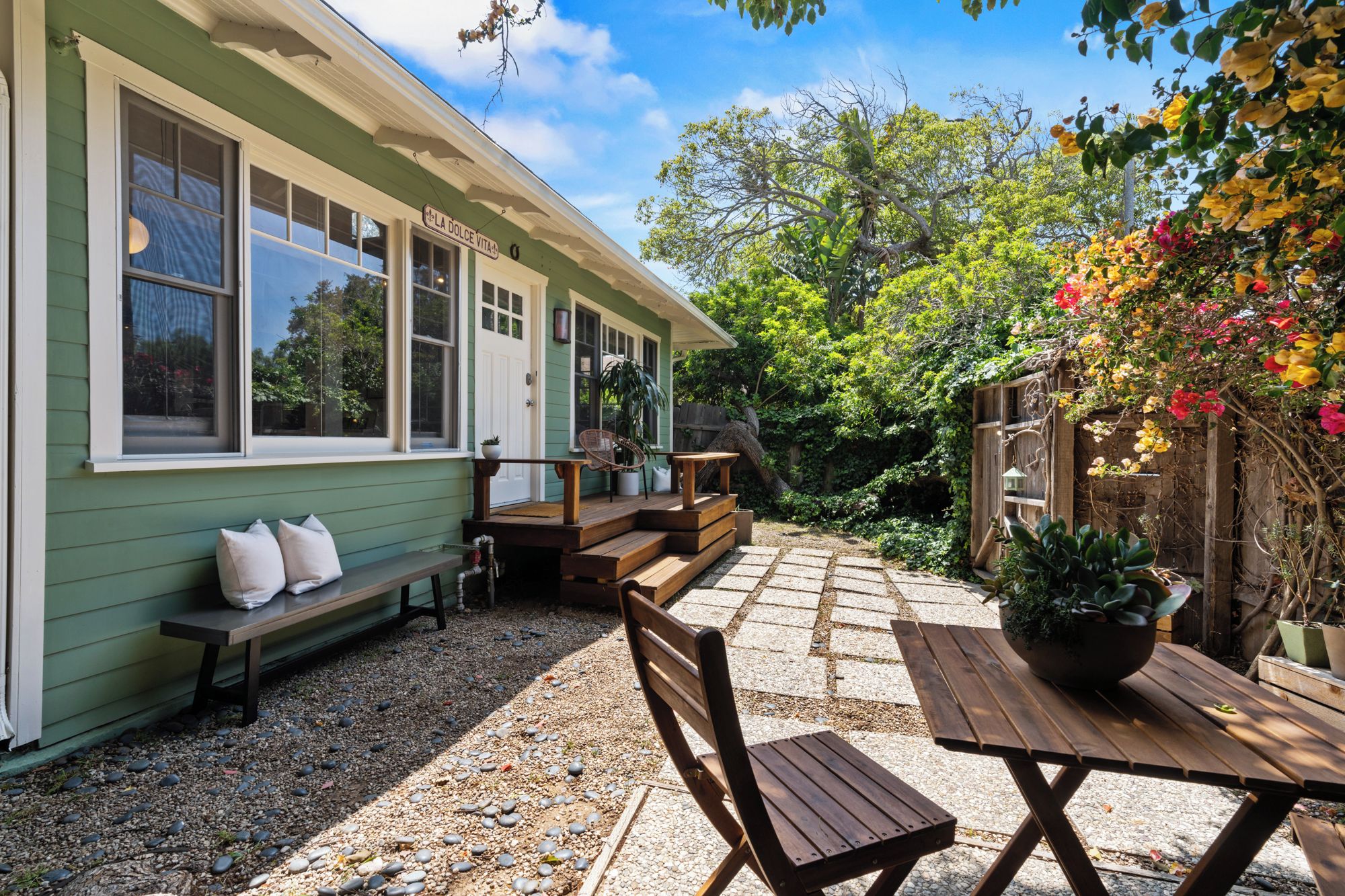 A house with a porch, a wooden bench, a table, and a garden with various plants.