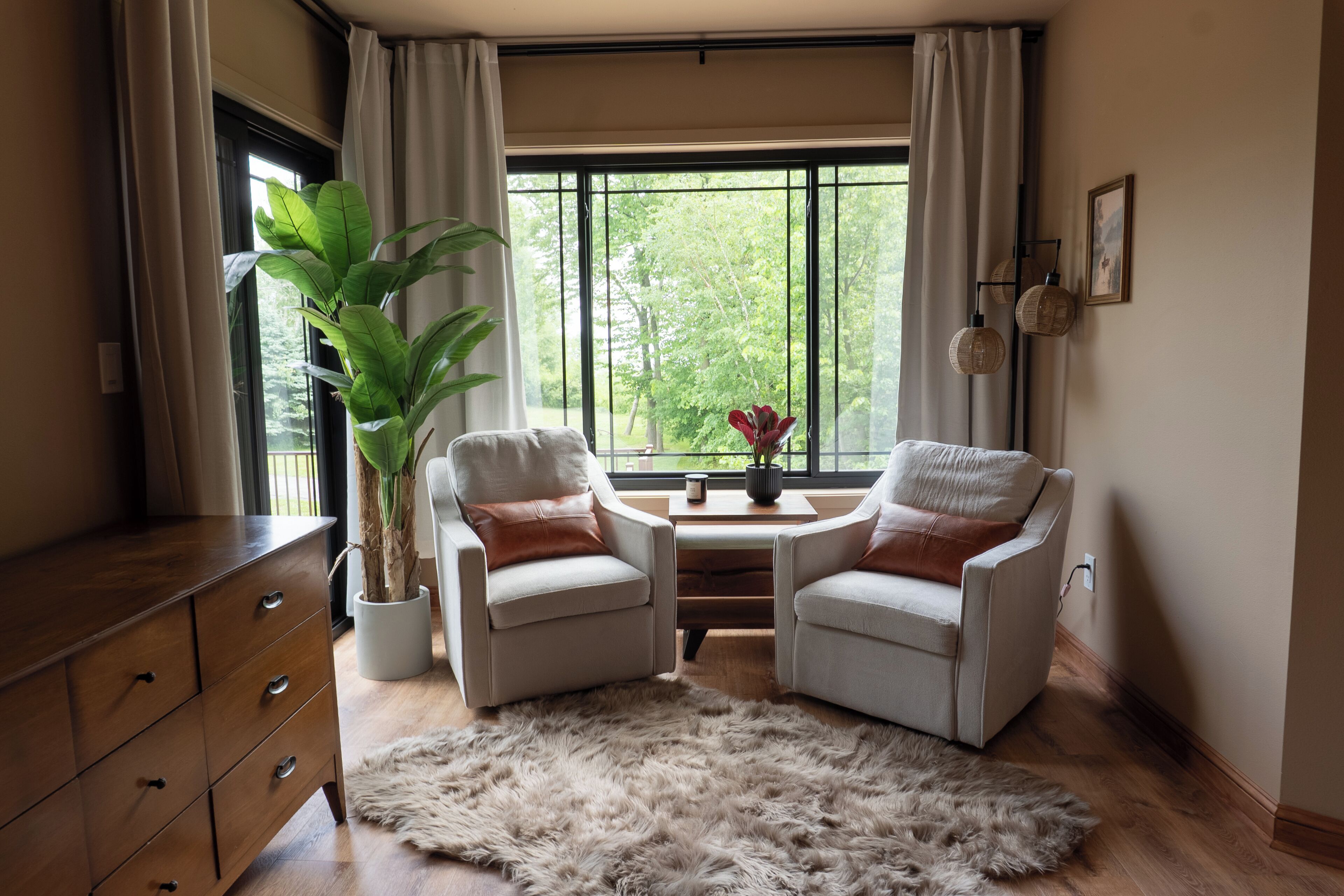 Une pièce comprenant deux fauteuils, une commode en bois, une grande plante en pot, une petite table avec un vase et une vue sur la verdure à travers de grandes fenêtres.
