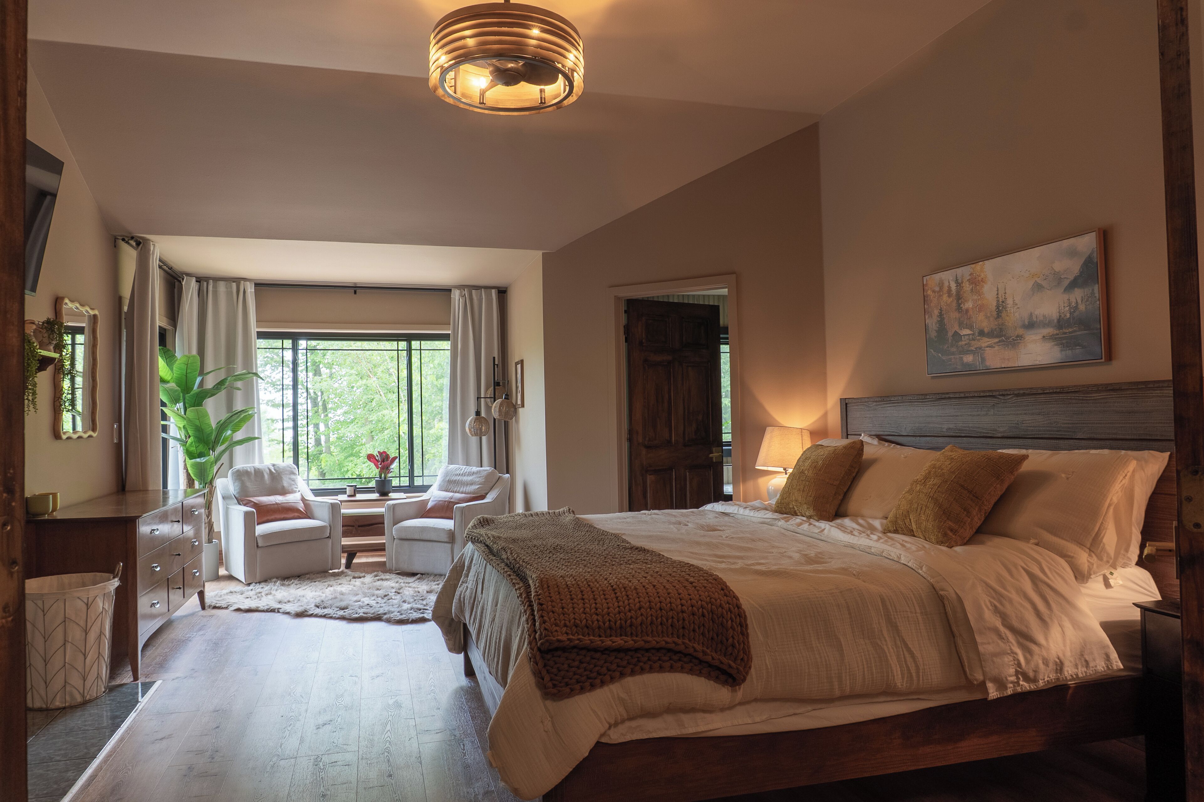 A bedroom with a large bed, a desk, two chairs, a window with curtains, and a painting on the wall.