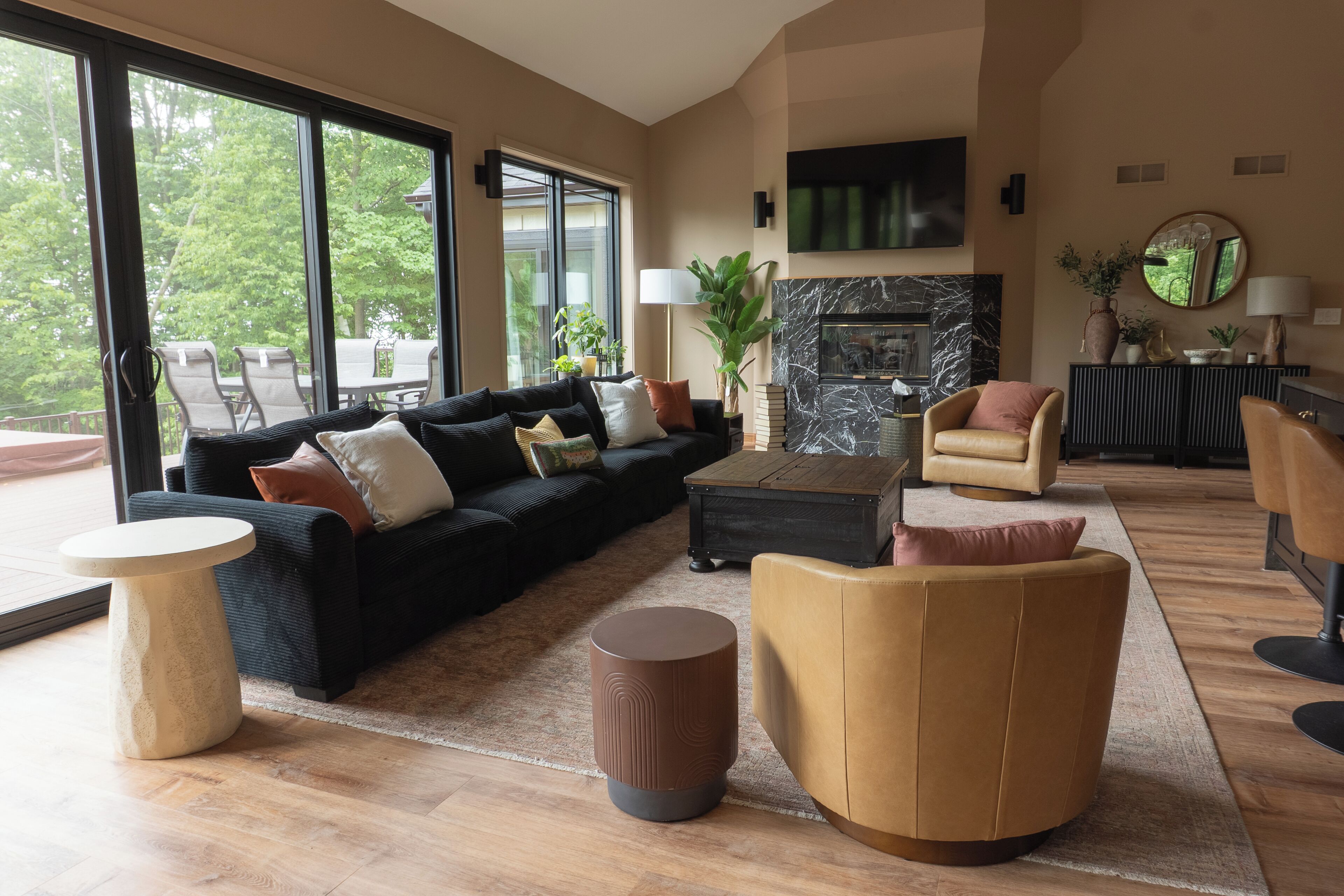 A spacious living room with a large black sofa, a round coffee table, and a beige armchair. There is a fireplace with a marble surround, a television mounted on the wall, and a large window providing a view of the outdoors.