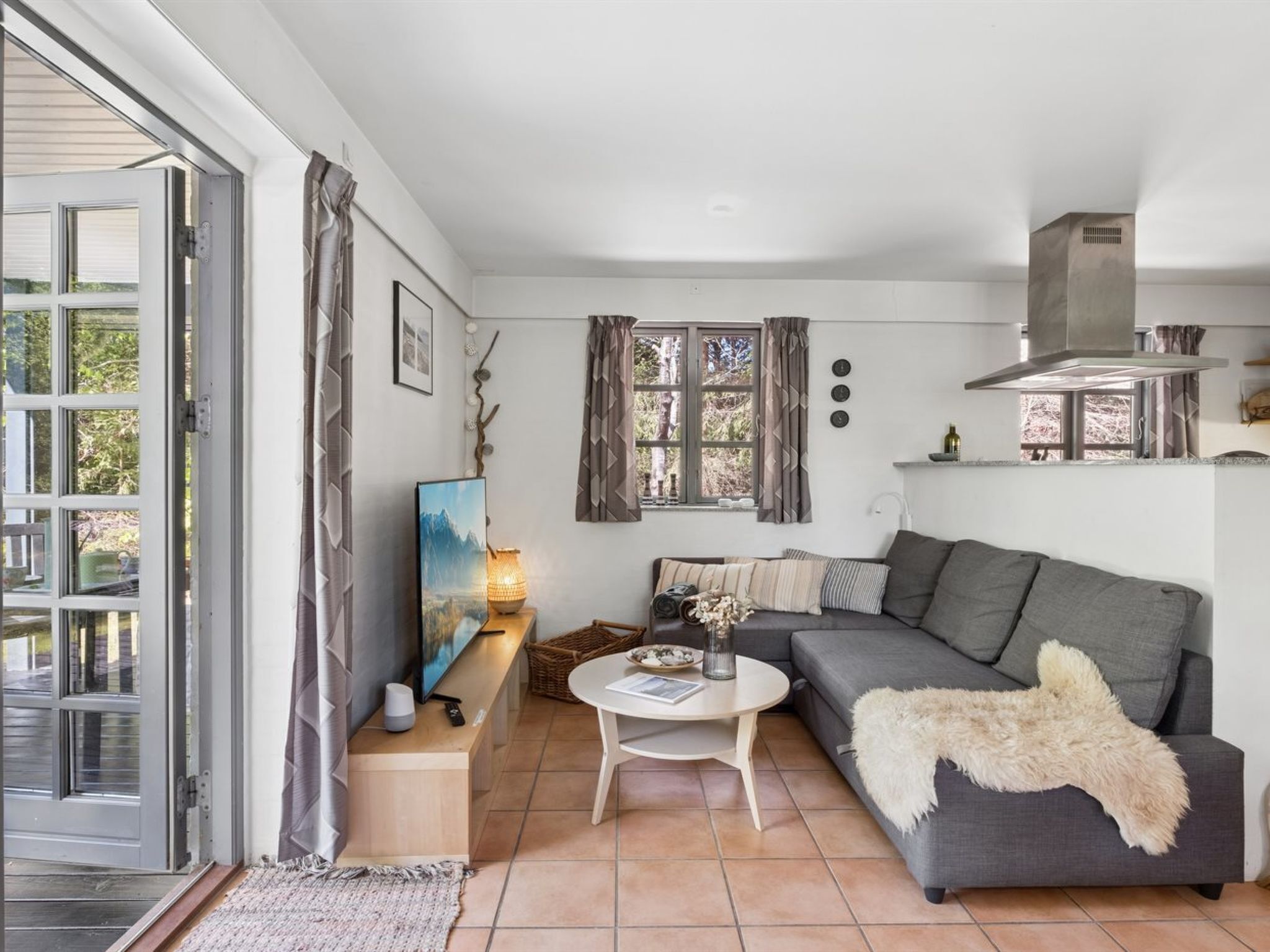 Uma sala de estar moderna com um sofá cinzento, uma mesa de centro branca e arredondada, uma TV sobre um suporte de madeira e uma janela grande com cortinas.