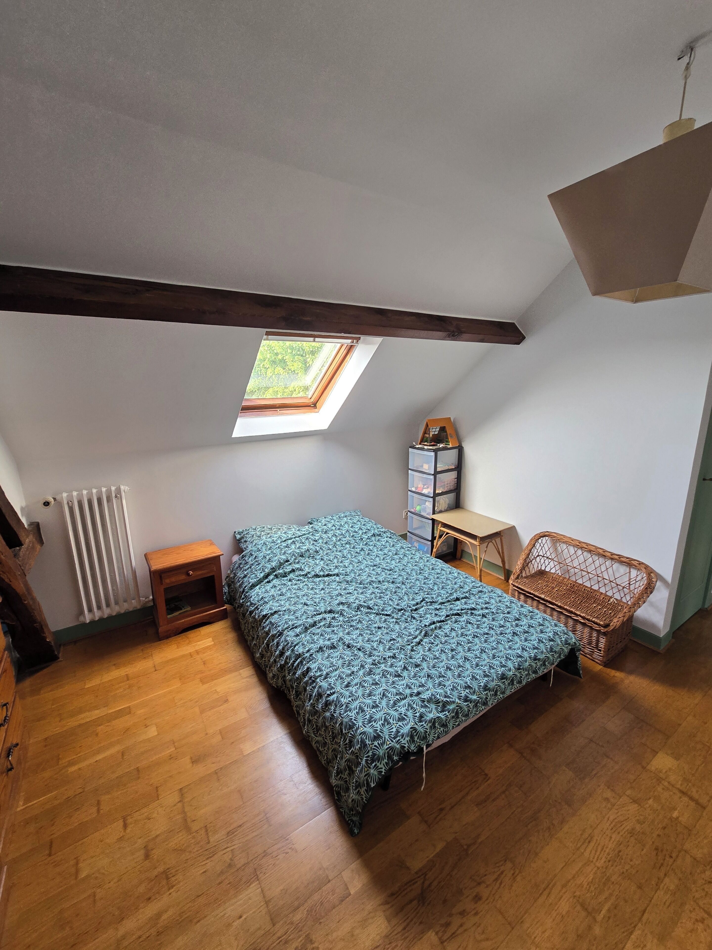 Ein Schlafzimmer mit schrägen Wänden, einem Dachfenster, einem Bett, einem kleinen Tisch, einem Korbstuhl und einer Heizung.