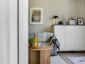 Ein Wohnzimmer mit einer Couch, einem runden Holztisch mit Flasche und Gläsern, einer Lampe und einer Kommode mit einem Bild und Kerzen.