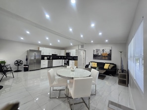 Eine moderne Küche mit weißen Schränken, ein Essbereich mit weißem Tisch und Stühlen und ein schwarzes Sofa mit gelben Kissen.