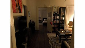 Una sala de estar con televisión, una estantería, un tapete estampado y una puerta que da a un dormitorio.