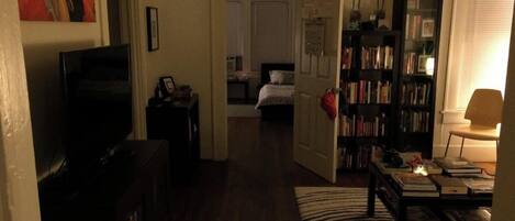 Una sala de estar con televisión, una estantería, un tapete estampado y una puerta que da a un dormitorio.