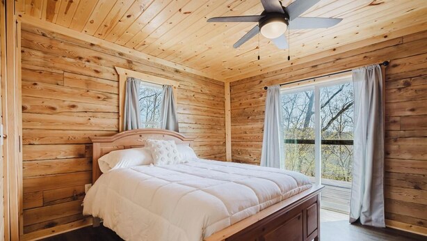 Ein Schlafzimmer einer Holzhütte mit Bett, Deckenventilator und Blick durch ein Fenster.