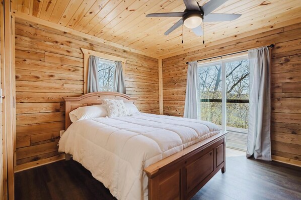 Ein Schlafzimmer einer Holzhütte mit Bett, Deckenventilator und Blick durch ein Fenster.