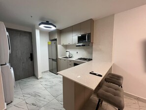 A compact kitchen with a microwave, stove, and sink, a small bar stool, and a door leading to another room. - Ocean View Apartment | High Floor | The Sands (Panamá)