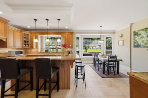 A kitchen with wooden cabinets, a central island with stools, pendant lighting, and a view to the outdoors. - Spacious 4-bedroom house in delightful North Vancouver with AC (North Vancouver)