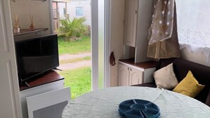 Un espacio habitable compacto con un televisor sobre un mueble, una mesa de comedor con mantel verde, un sofá con cojines y un puerta corredera de vidrio que da a un área exterior.