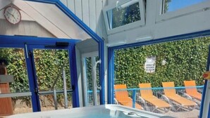 Un jacuzzi con revestimiento de madera, rodeado por una estructura azul y blanca, y una vista de sillones anaranjados y follaje al fondo.